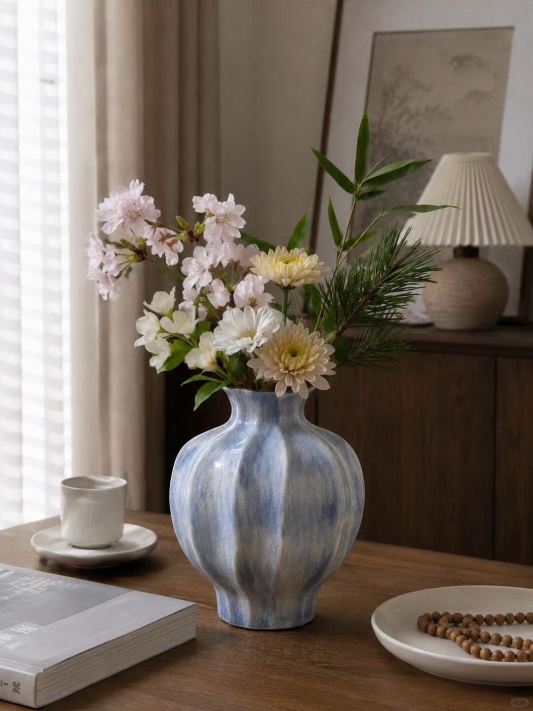 Pale pink, white, and cream flowers with green branches fill a blue-and-white lobed vase on a wooden table, beside a cup, book, and dish of wooden beads. Curtains, a cabinet, framed artwork, and a pleated-shade lamp form the background.