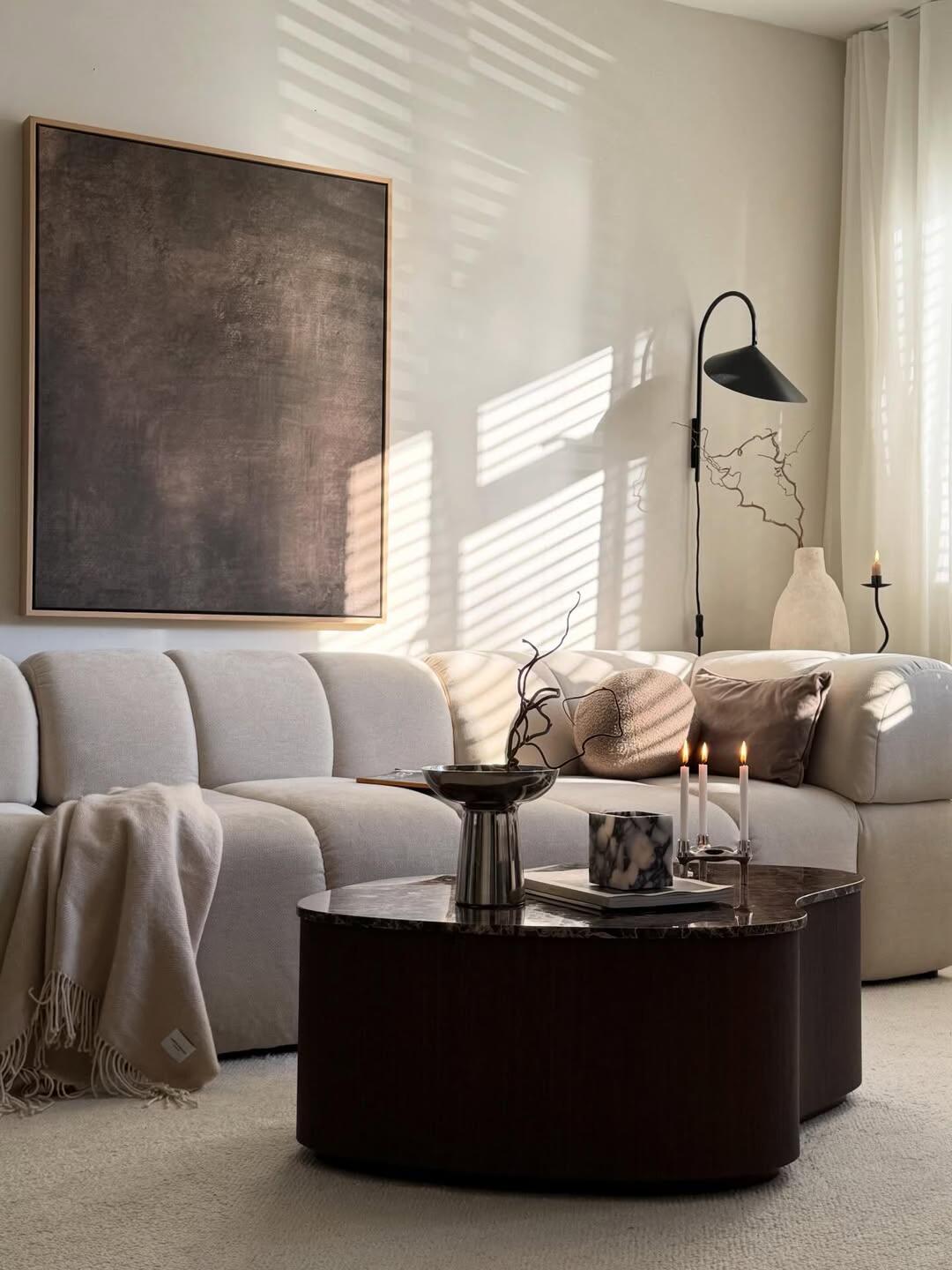 Sunlit neutral-toned living room with a cream sectional, large dark abstract painting, and rounded dark coffee table displaying branches, books, and lit candles. Striped shadows cross the wall and sofa beside white curtains.