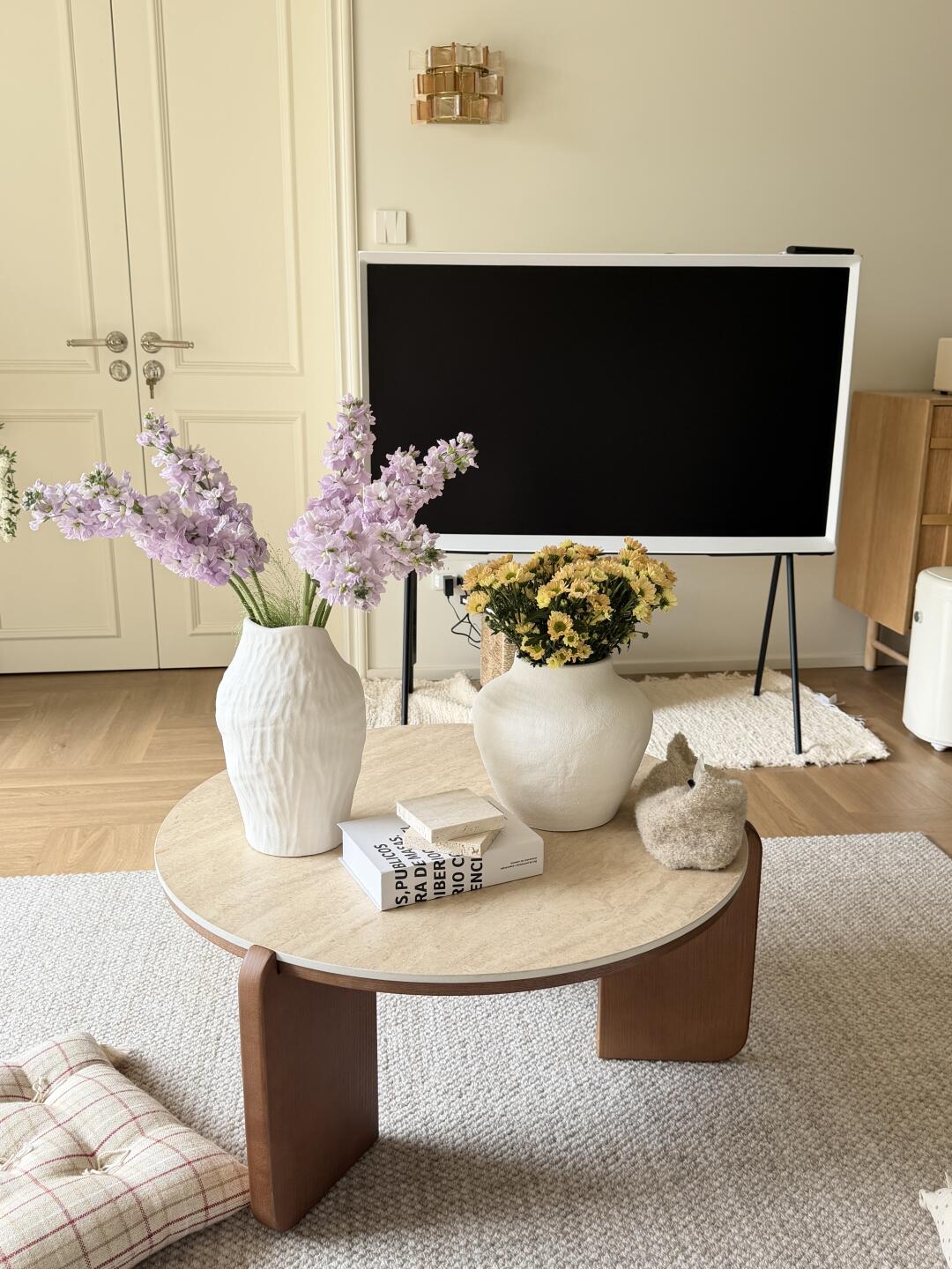 Neutral-toned living room with lavender and yellow flowers in white vases on a rounded coffee table, a black-screen television, cream double doors, and textured rugs.