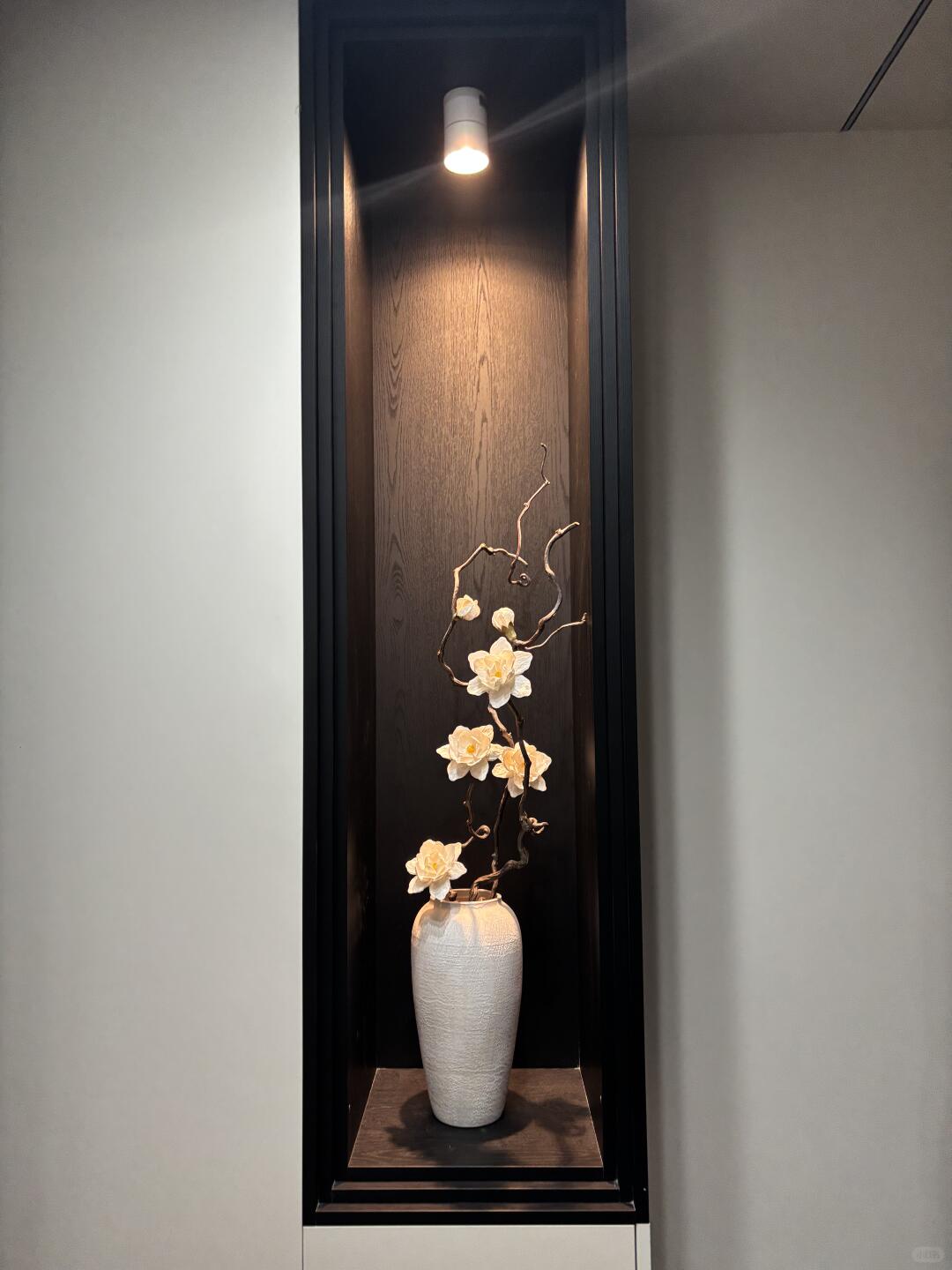 An illuminated dark wood wall niche holds a tall off-white vase with twisting branches and cream-colored flowers, framed by black trim and pale gray walls.