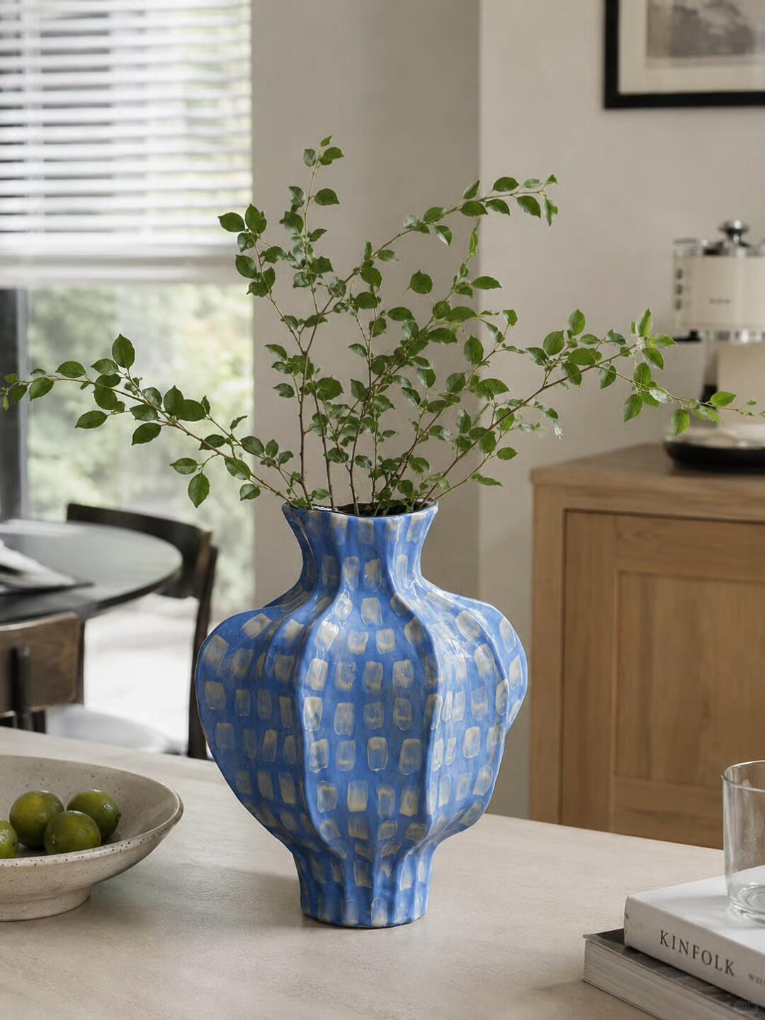 Sculptural blue vase with cream rectangular marks and spreading leafy branches on a pale countertop, beside a bowl of green citrus and stacked books.