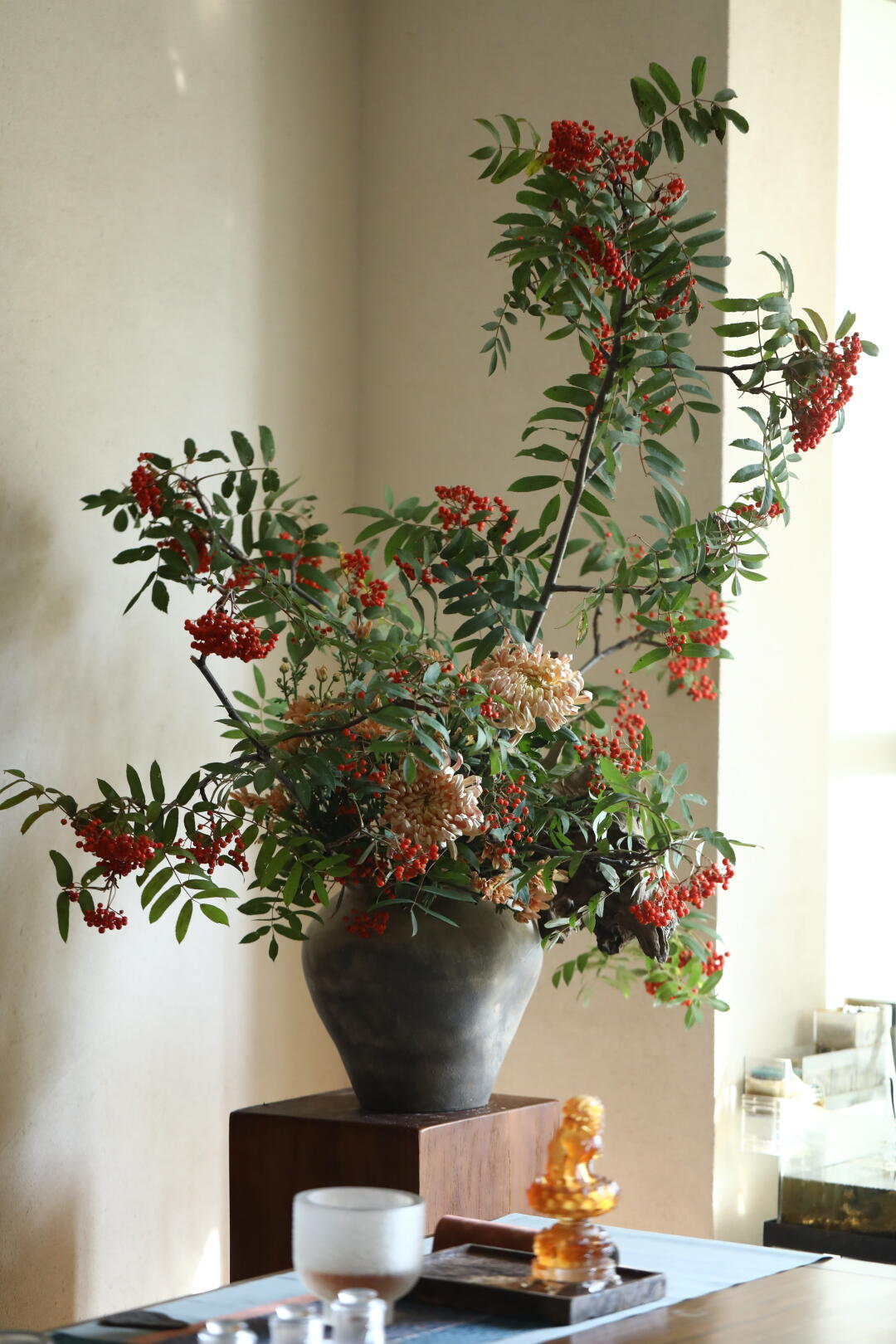 Tall branches of red-orange berries and green leaves, with pale peach flowers, arranged in a charcoal-gray vase on a wooden pedestal beside a sunlit wall. A cup and amber-colored figurine sit on a foreground table.