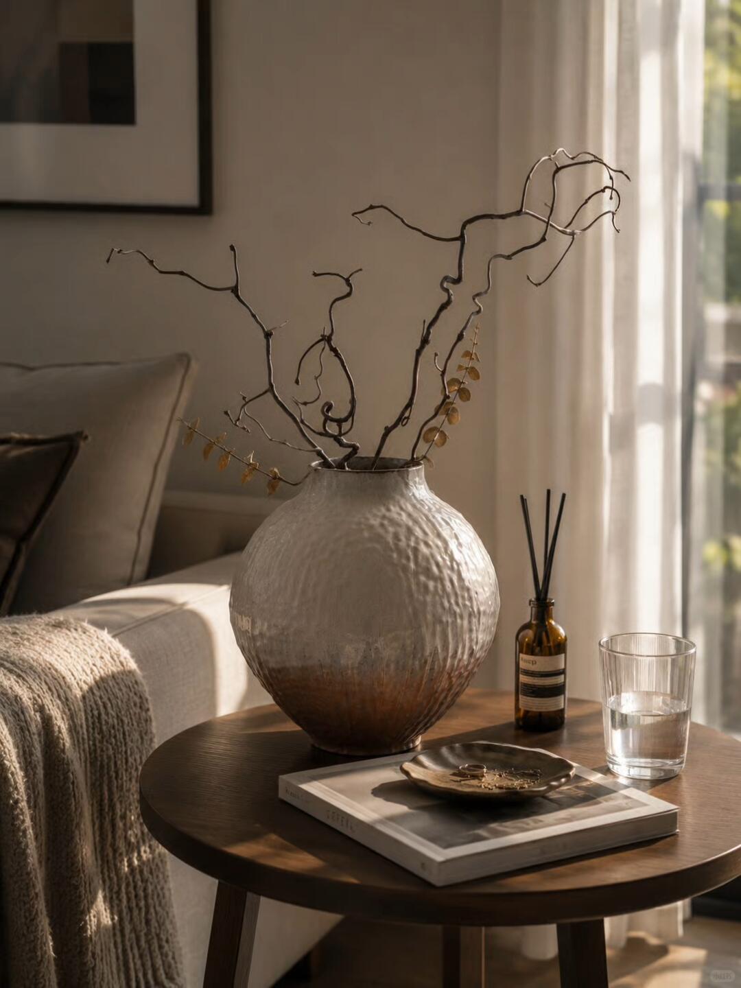 Sunlit wooden side table with a large textured vase of twisting branches, a reed diffuser, a glass of water, and a jewelry dish on a book, beside a sofa with a knitted throw.