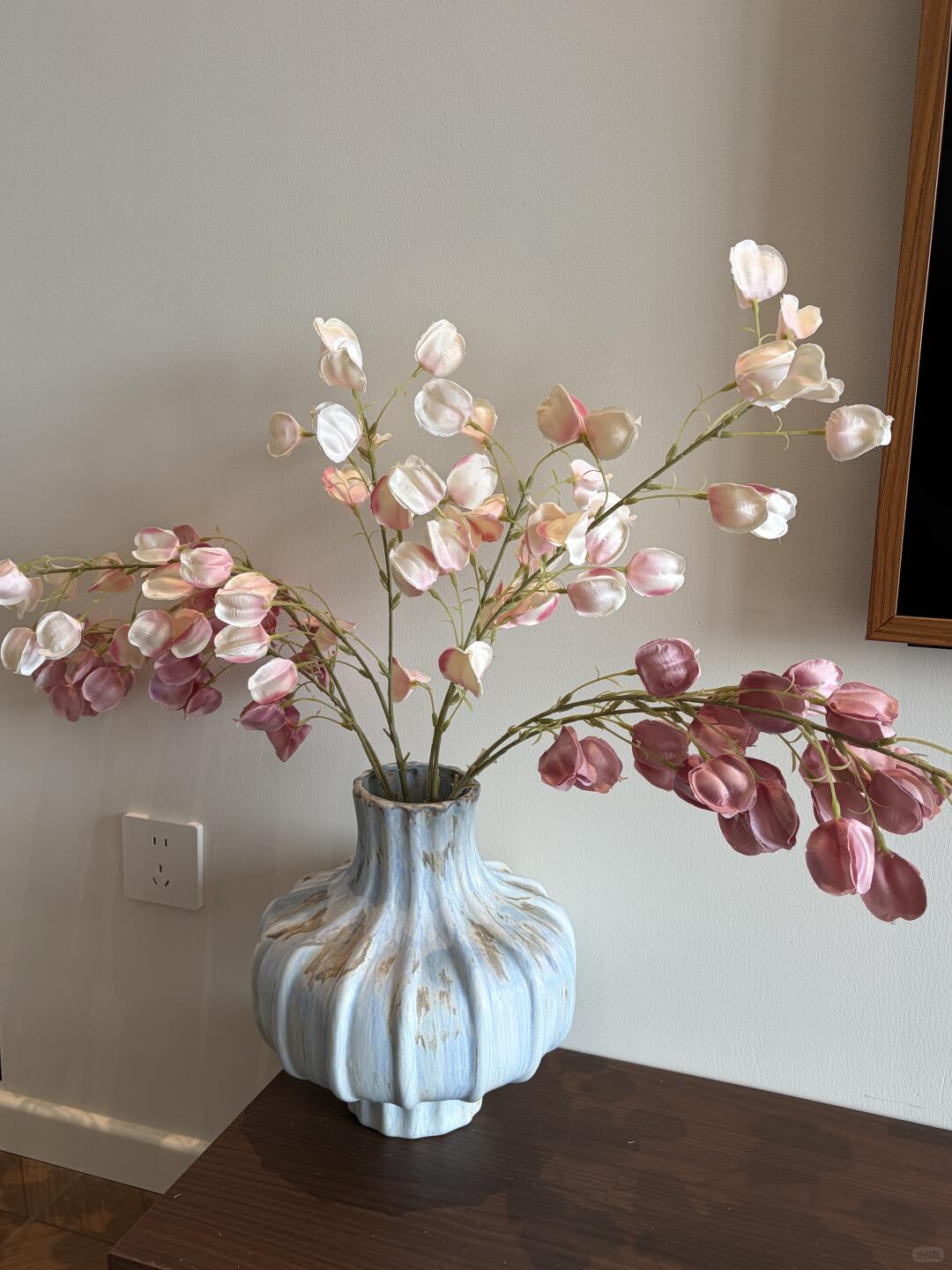 Pale pink, ivory, and mauve flower stems spread from a ribbed pale blue vase on a dark wooden table against a warm gray wall.