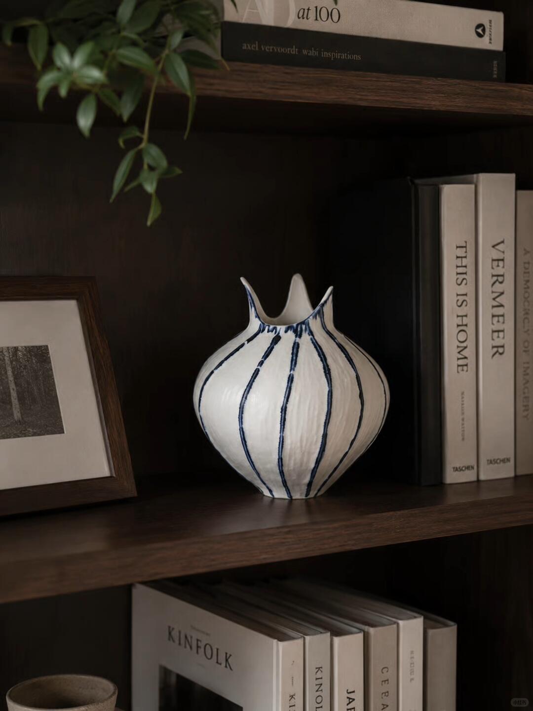 Dark wooden shelves with a white vase painted in blue vertical stripes, a framed woodland picture, pale art books, and trailing green leaves.